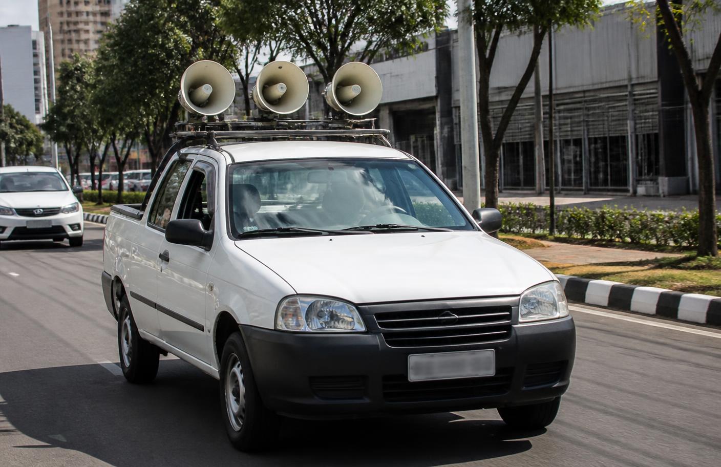 Carro de som com alto-falantes trafegando em avenida urbana durante o dia